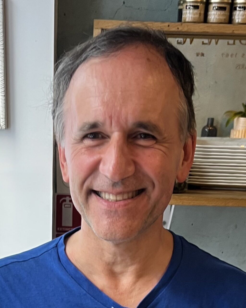 A smiling middle-aged man with short, graying hair wears a blue shirt and stands indoors with shelves, plates, and plants visible in the background.