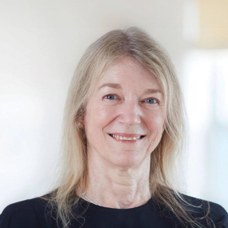 Smiling older woman with long, light blonde hair, wearing a black top, facing the camera against a soft, light background.