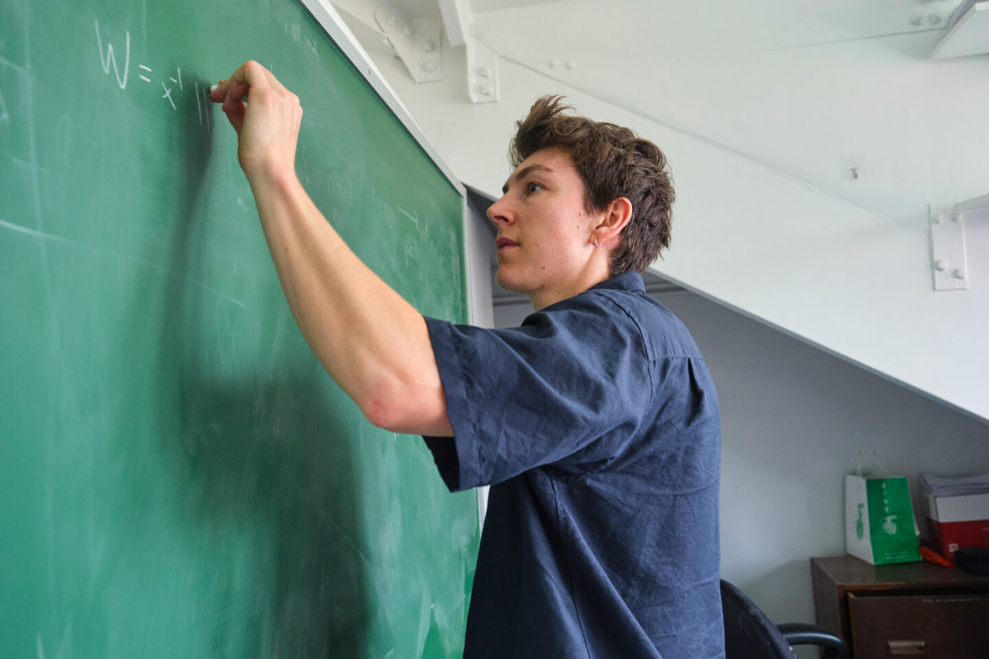 A person writes a mathematical equation on a green chalkboard in a classroom, wearing a dark shirt and looking focused.