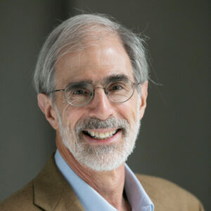 Smiling older man with white hair, beard, and glasses, wearing a light blue shirt and brown blazer, standing in front of a dark, neutral background.