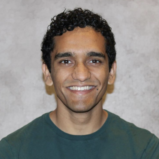 A person with short curly hair and medium skin tone, wearing a dark green shirt, is smiling at the camera in front of a light gray textured background.