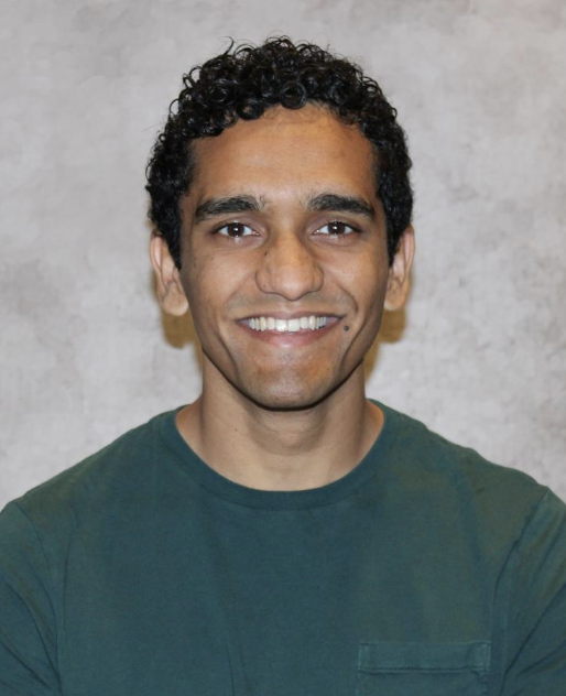 A person with short curly hair and medium skin tone, wearing a dark green shirt, is smiling at the camera in front of a light gray textured background.