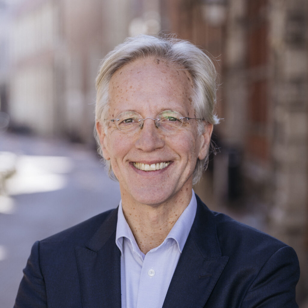 An older man with gray hair and glasses smiles while standing outside in a city street, wearing a navy suit and light blue shirt.
