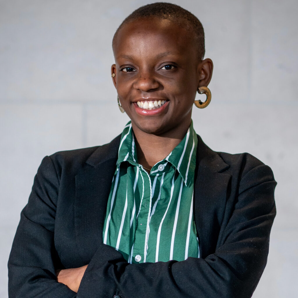A person with short hair, wearing a black blazer over a green and white striped shirt, smiles confidently with arms crossed against a plain light background.