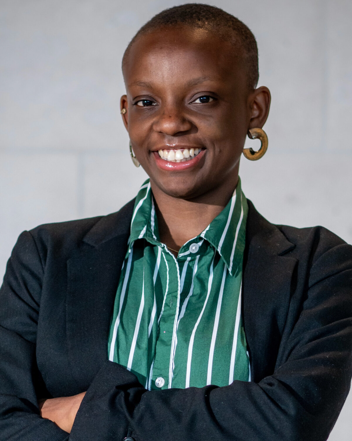A person with short hair, wearing a black blazer over a green and white striped shirt, smiles confidently with arms crossed against a plain light background.
