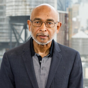A man with glasses, a grey beard, and a bald head, wearing a dark blazer and patterned shirt, stands in front of a window with an urban background.