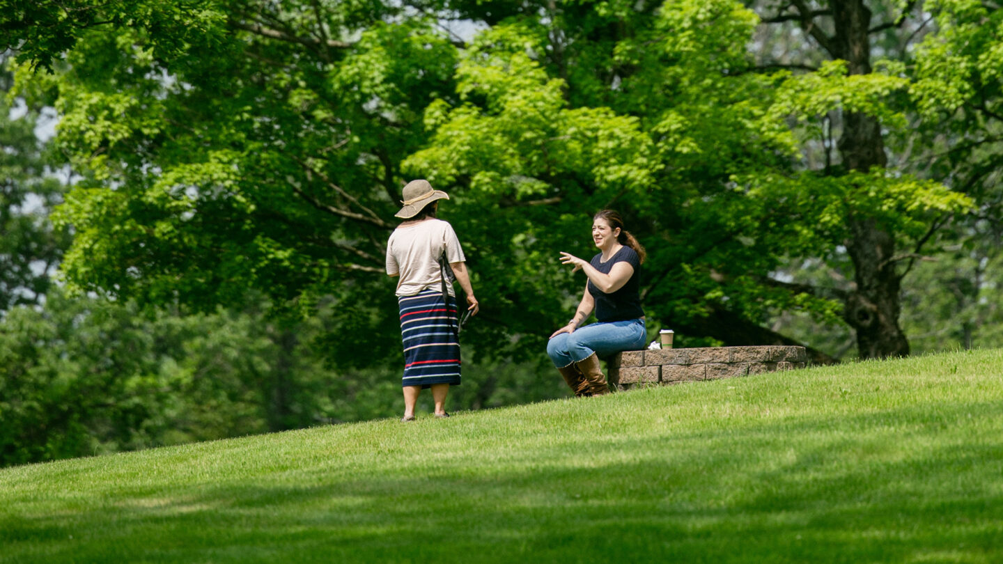 Two people interact on a grassy hill; one is standing wearing a hat and striped skirt, while the other sits on a stone ledge gesturing, surrounded by green trees in the background.