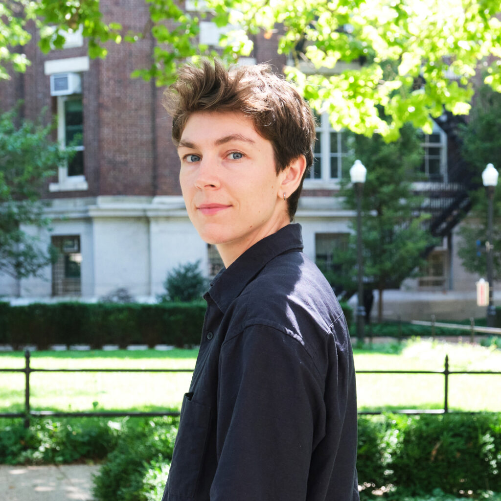 A person with short brown hair and a dark shirt stands outdoors in a sunny park, with green trees, grass, and a brick building in the background.