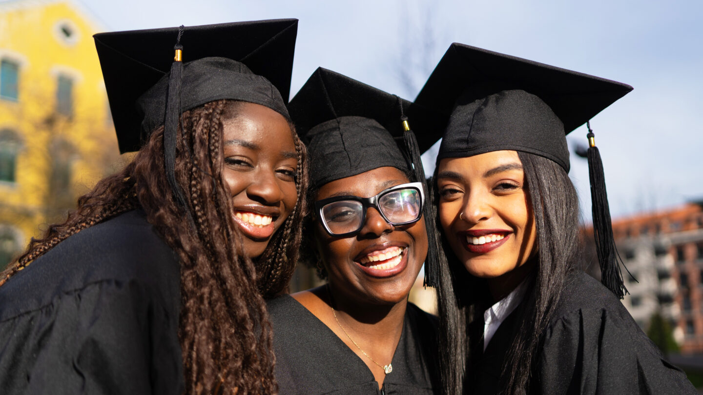Three graduates in black caps and gowns smile closely together outdoors, celebrating their achievement. A building and trees are blurred in the background under a sunny sky.