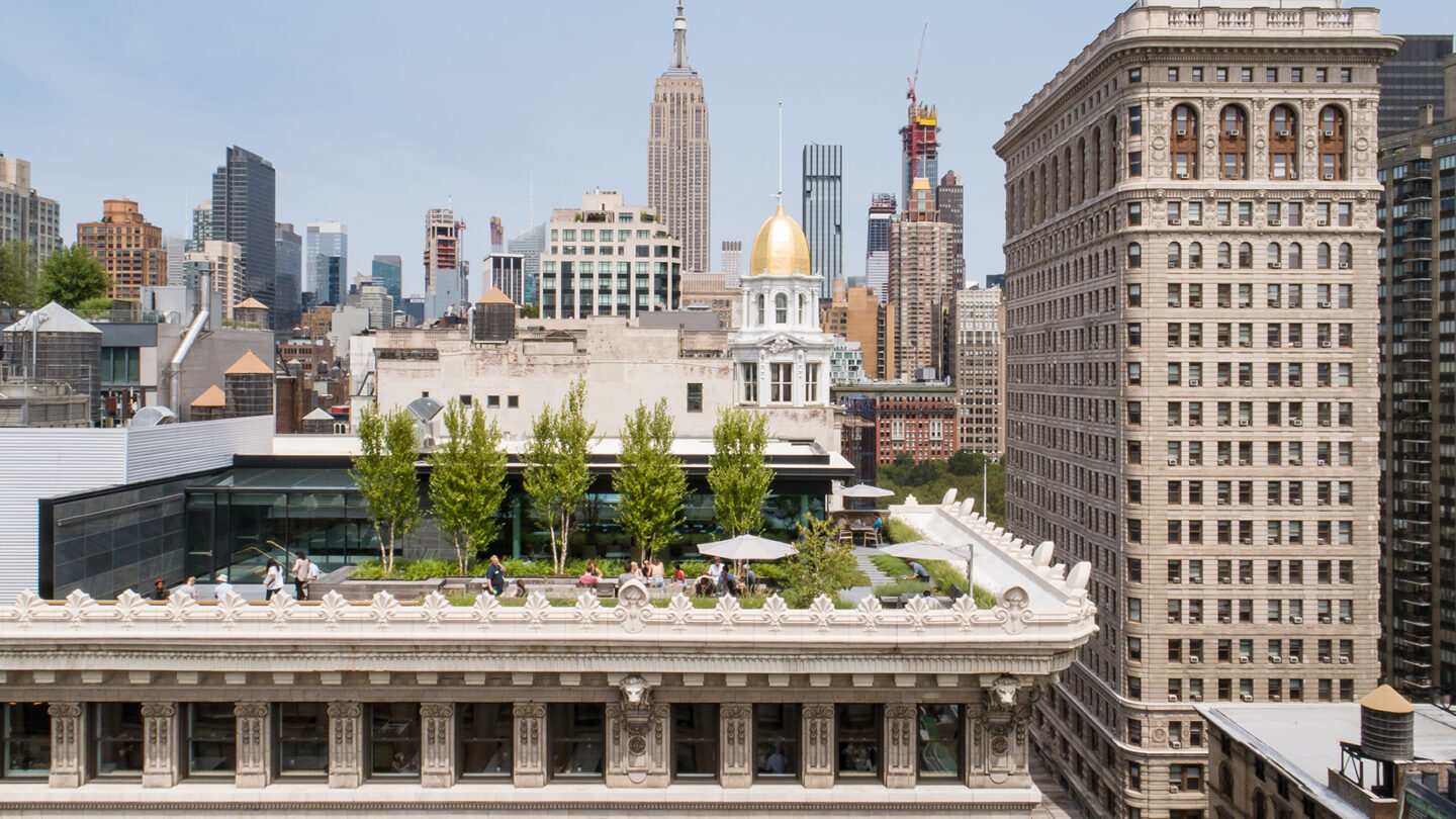 A rooftop garden with people relaxing and trees, set against a backdrop of New York City skyscrapers, including the Empire State Building and the Flatiron Building, under a clear sky.