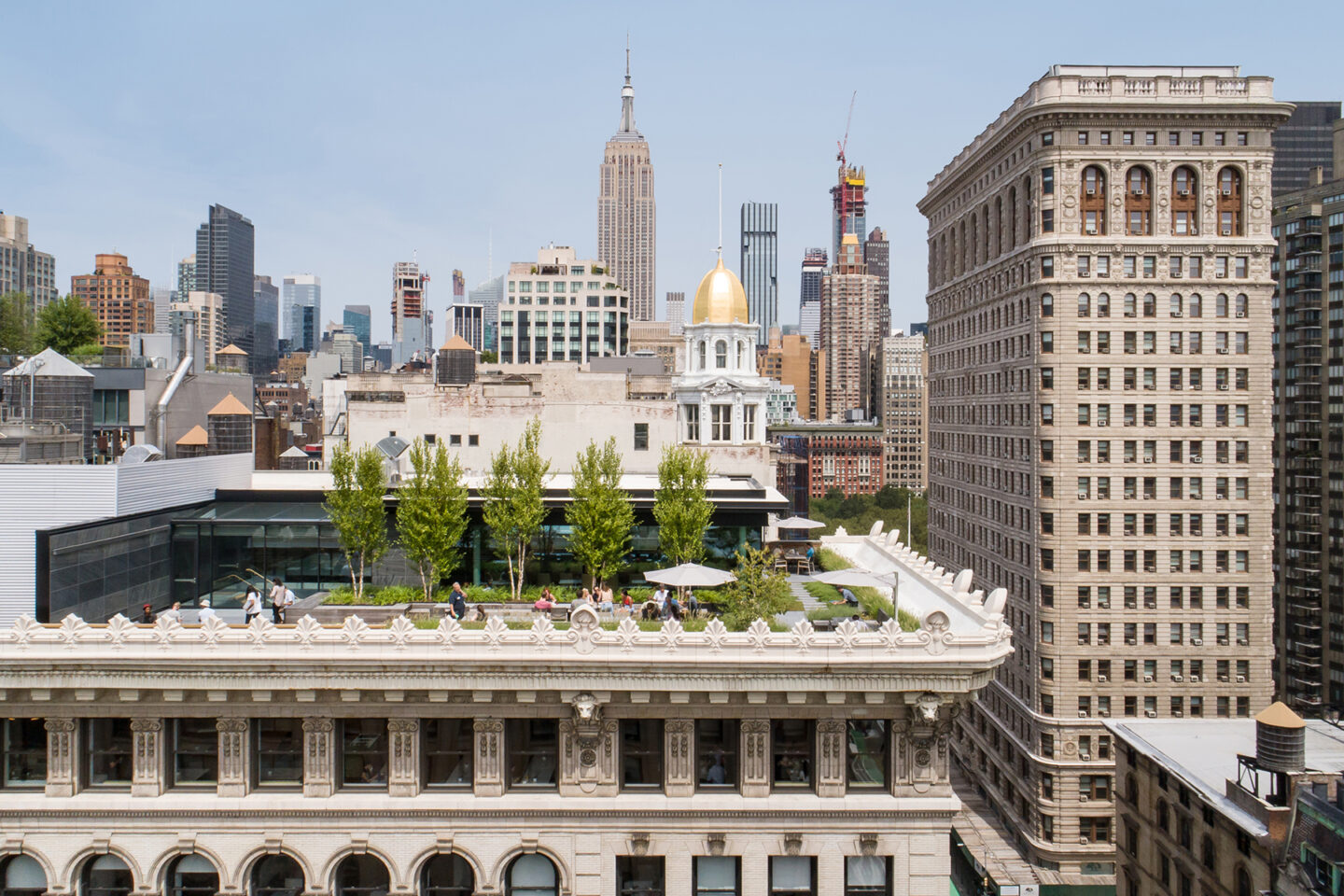 A rooftop garden with people relaxing and trees, set against a backdrop of New York City skyscrapers, including the Empire State Building and the Flatiron Building, under a clear sky.