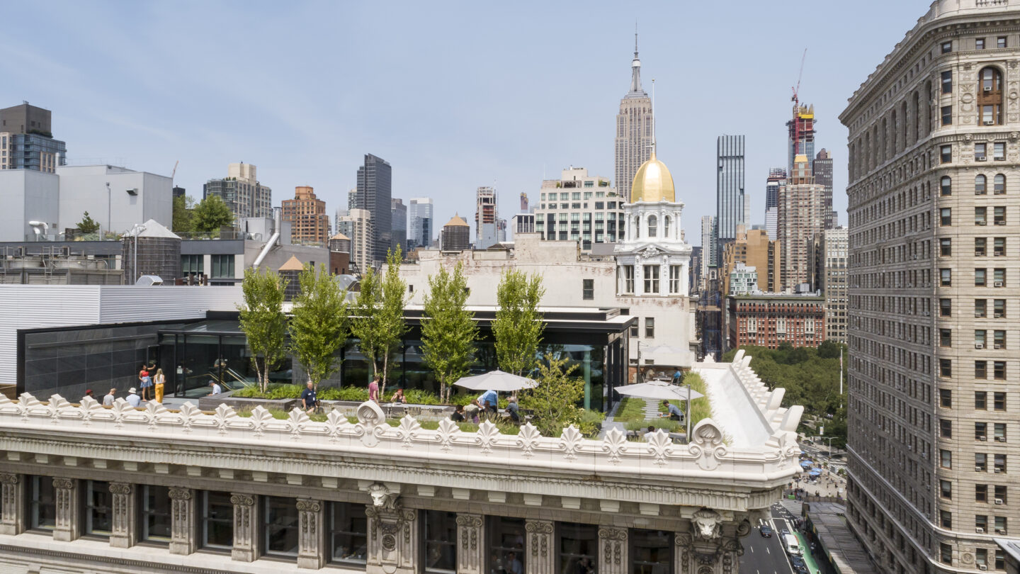 Rooftop garden with trees and people atop an ornate historic building in New York City, with skyscrapers and the Empire State Building visible in the background.