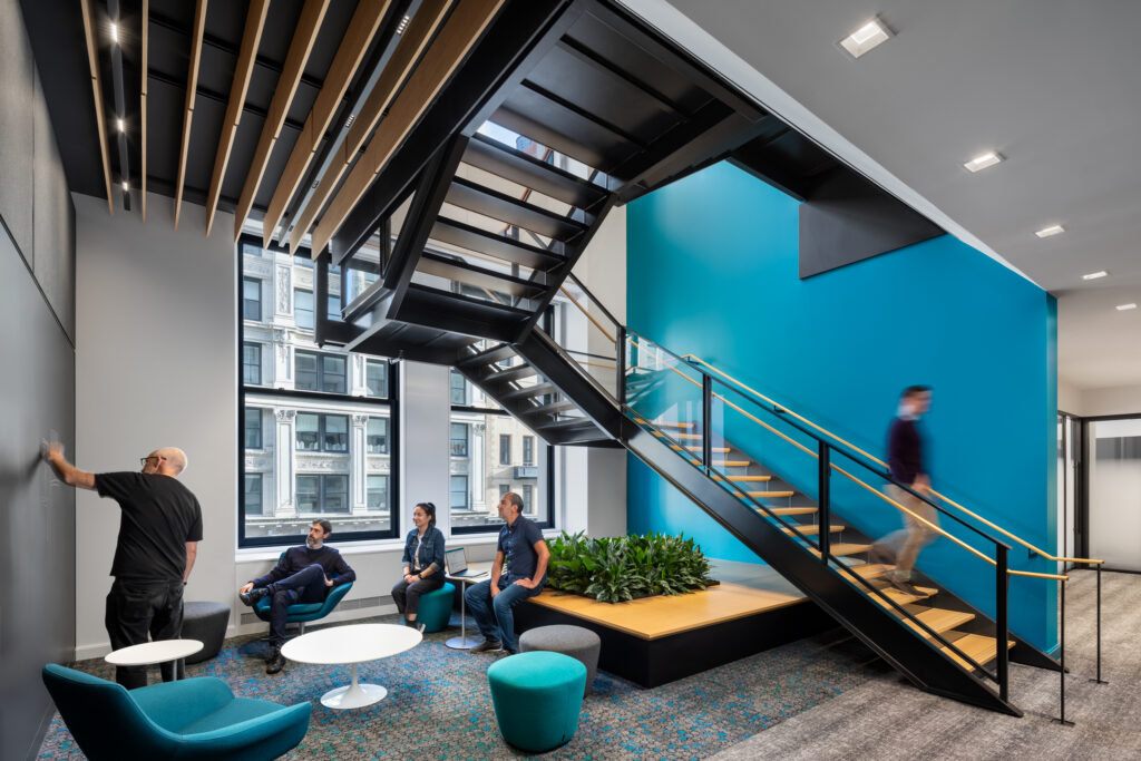 Modern office lounge with four people sitting and talking near round tables, one person writing on a wall, and another person walking up stairs next to a large window and blue accent wall.
