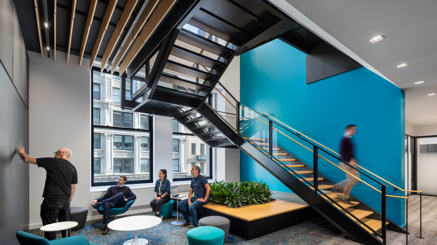 Modern office lounge with four people sitting and talking near round tables, one person writing on a wall, and another person walking up stairs next to a large window and blue accent wall.