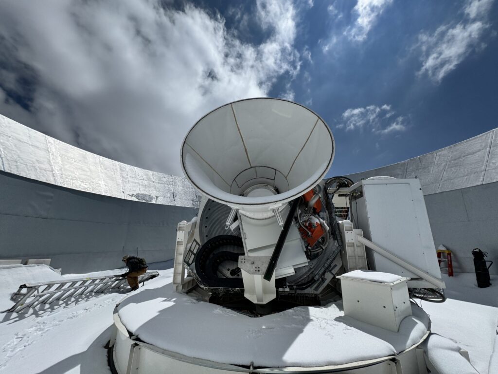 A large white radio telescope dish is positioned outdoors in a snowy observatory, surrounded by high walls under a partly cloudy sky. A metal ladder rests on the snow nearby.
