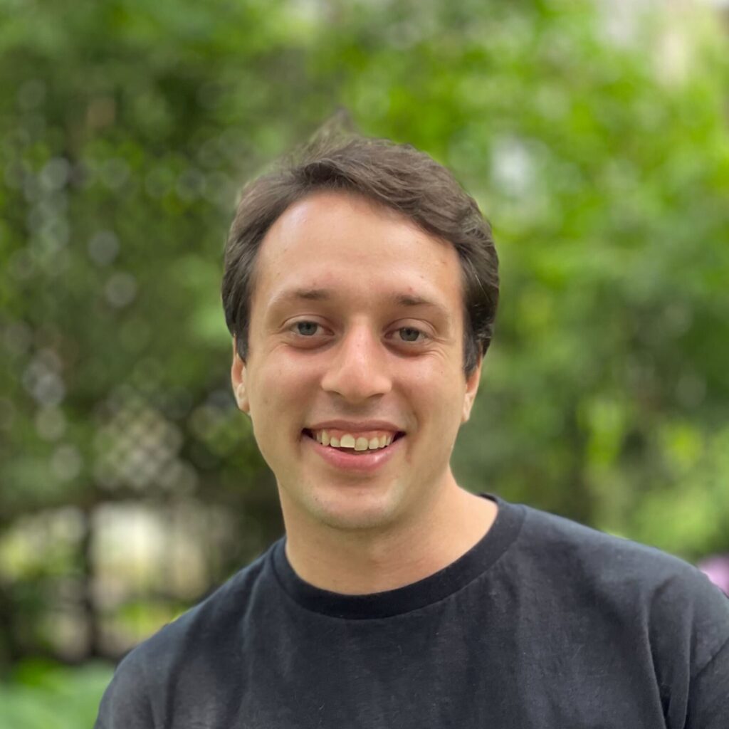 A person with short brown hair wearing a black t-shirt smiles at the camera outdoors, with green trees and blurred foliage in the background.