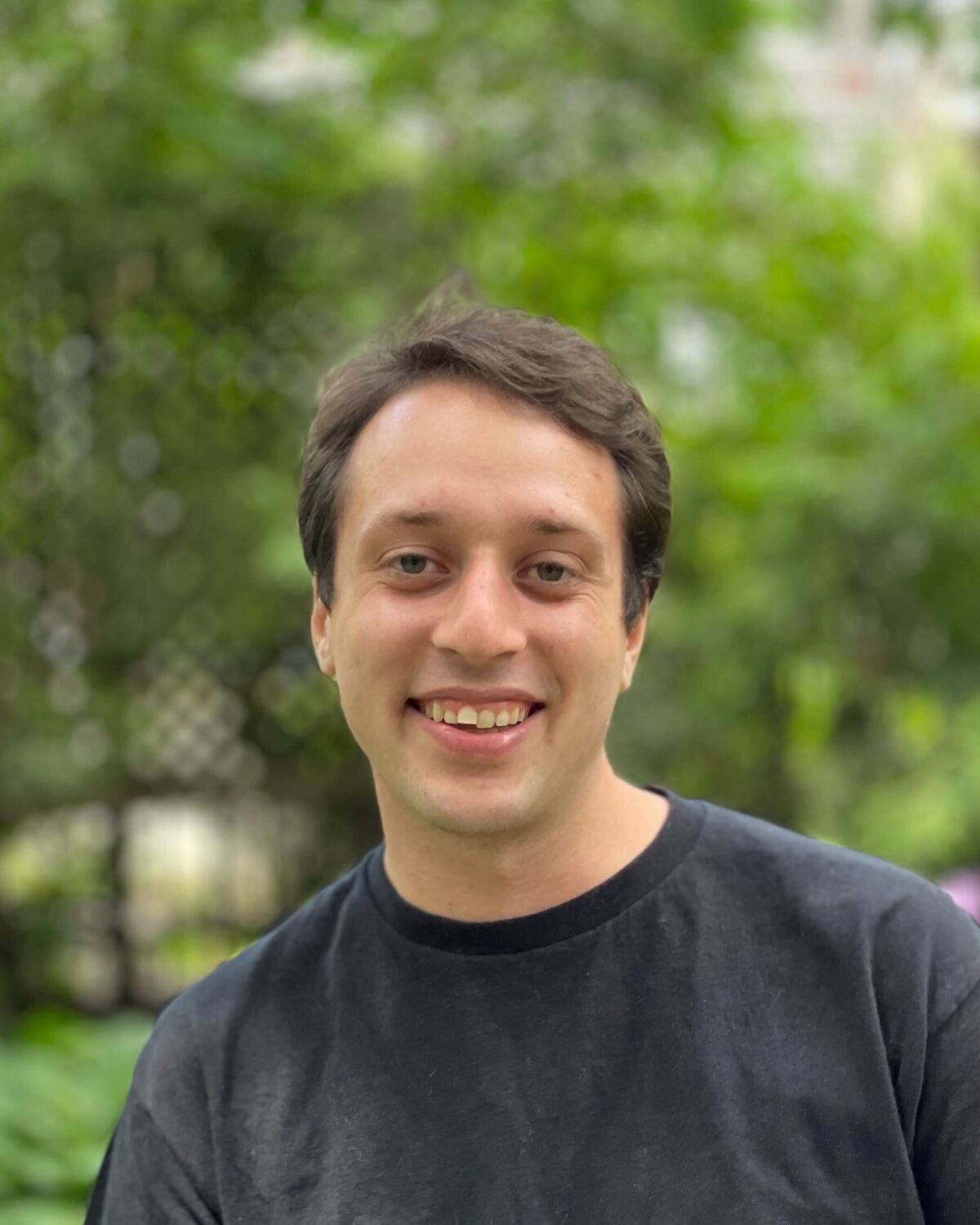 A person with short brown hair wearing a black t-shirt smiles at the camera outdoors, with green trees and blurred foliage in the background.