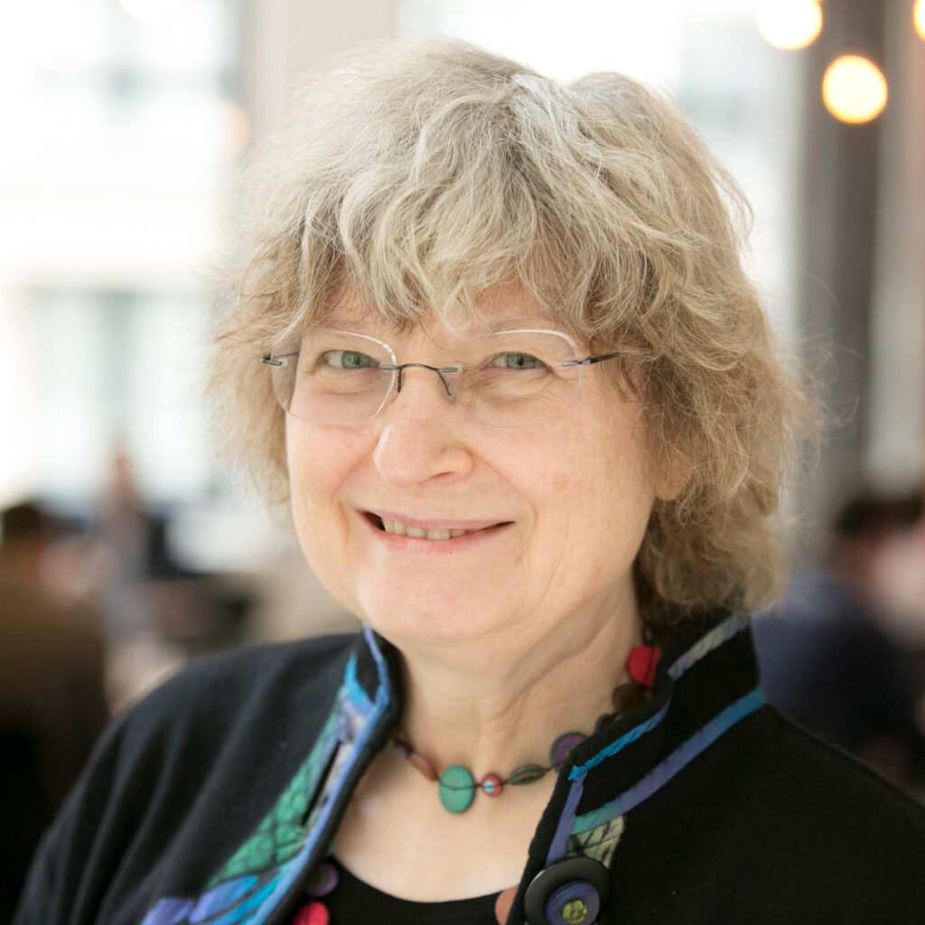 An older woman with curly gray hair, glasses, and a colorful beaded necklace smiles at the camera indoors with a blurred background.