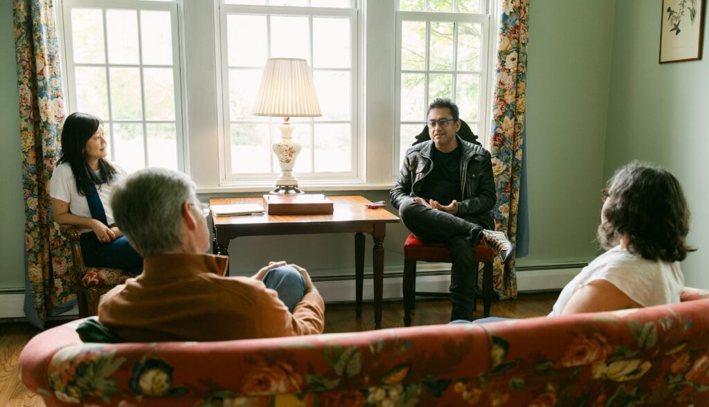 Four people are having a conversation in a cozy living room with floral curtains, a floral-patterned couch, and a lit lamp on a wooden table in front of a large window.