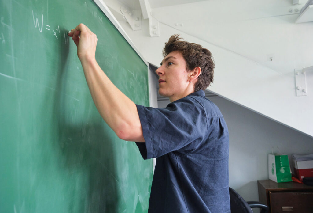 A person in a dark shirt writes a mathematical equation on a green chalkboard in a classroom, facing sideways.