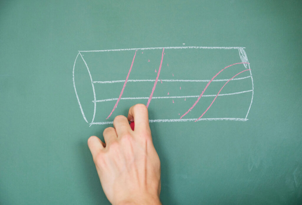 A hand draws a white chalk cylinder with red helical lines on a green chalkboard, illustrating a mathematical or scientific concept.