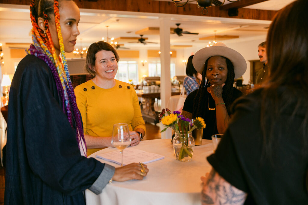 Four people sit around a round table with drinks and flowers, engaged in conversation. One person has colorful braids, another wears yellow, and another wears a hat. The setting looks warm and inviting.