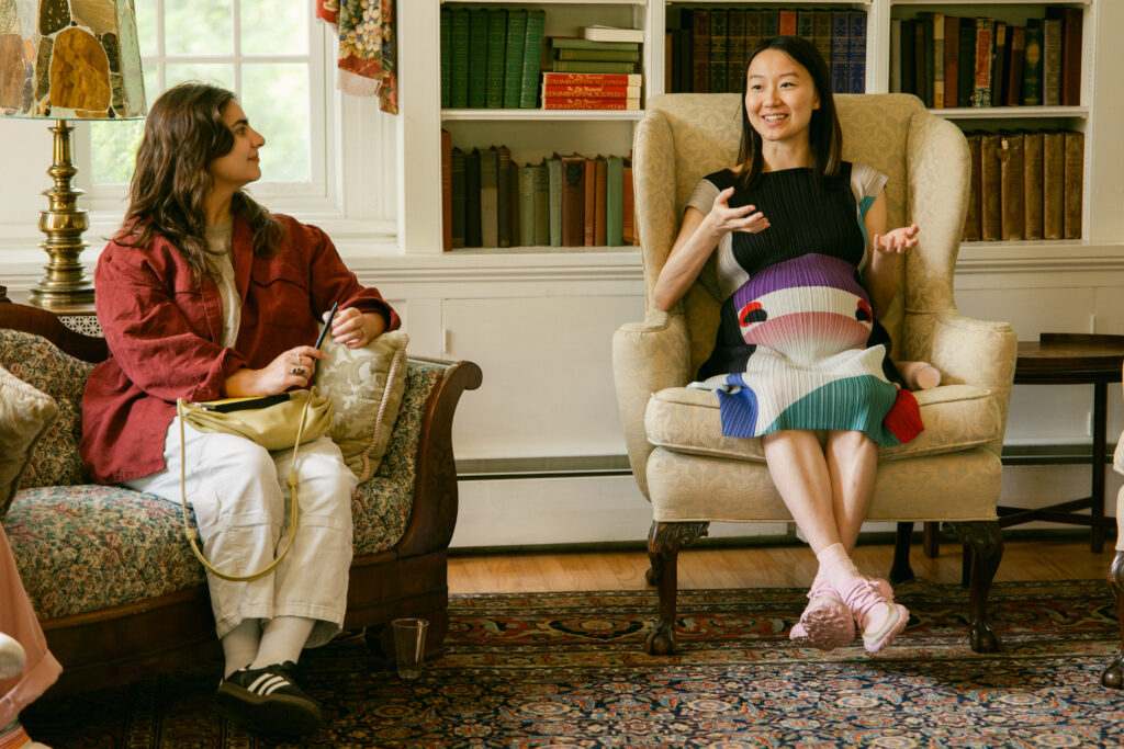 Two women sit in a cozy, book-filled room. One, wearing a colorful dress, gestures while speaking. The other, in a red jacket and white pants, listens attentively on a patterned sofa beside a window.