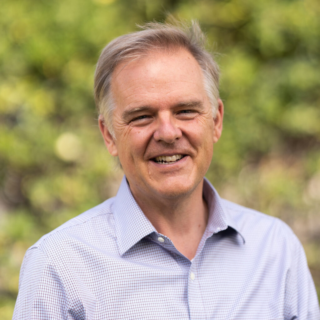 A middle-aged man with light hair smiles outdoors, wearing a light blue checkered shirt. The background is blurred greenery.