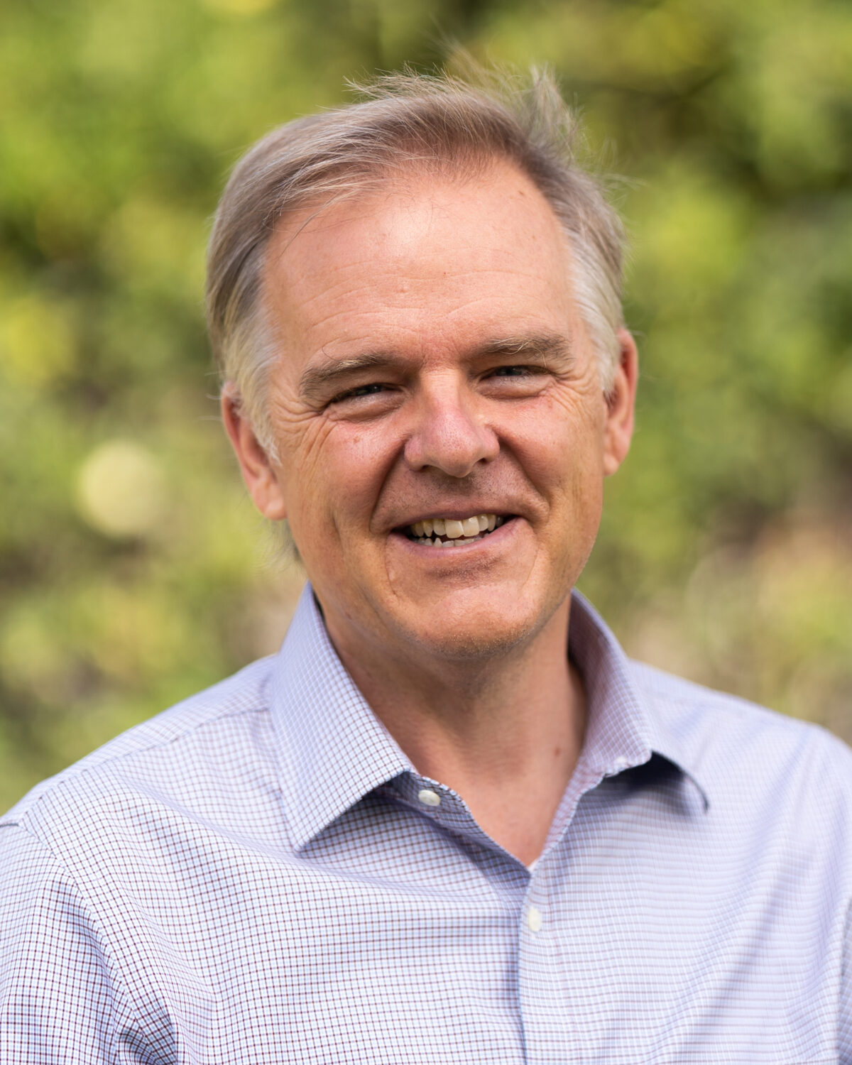 A middle-aged man with light hair smiles outdoors, wearing a light blue checkered shirt. The background is blurred greenery.