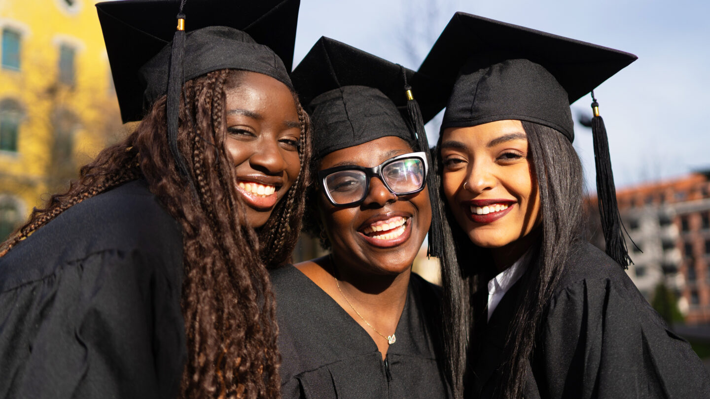 Three young women in black graduation caps and gowns stand close together, smiling and celebrating outdoors on a sunny day, with buildings blurred in the background.