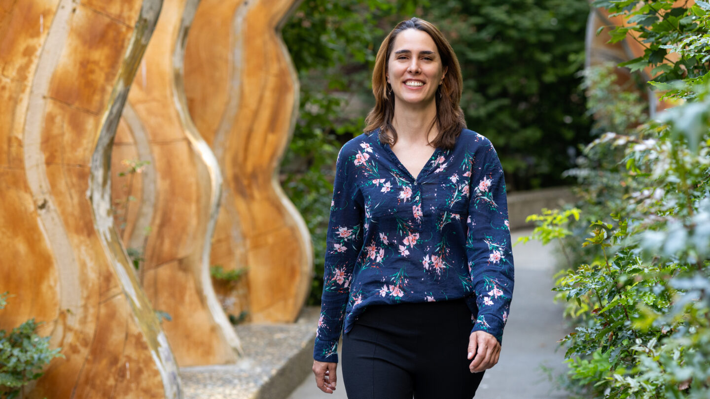 A smiling woman wearing a dark floral blouse walks along a path next to large, curved wooden sculptures.