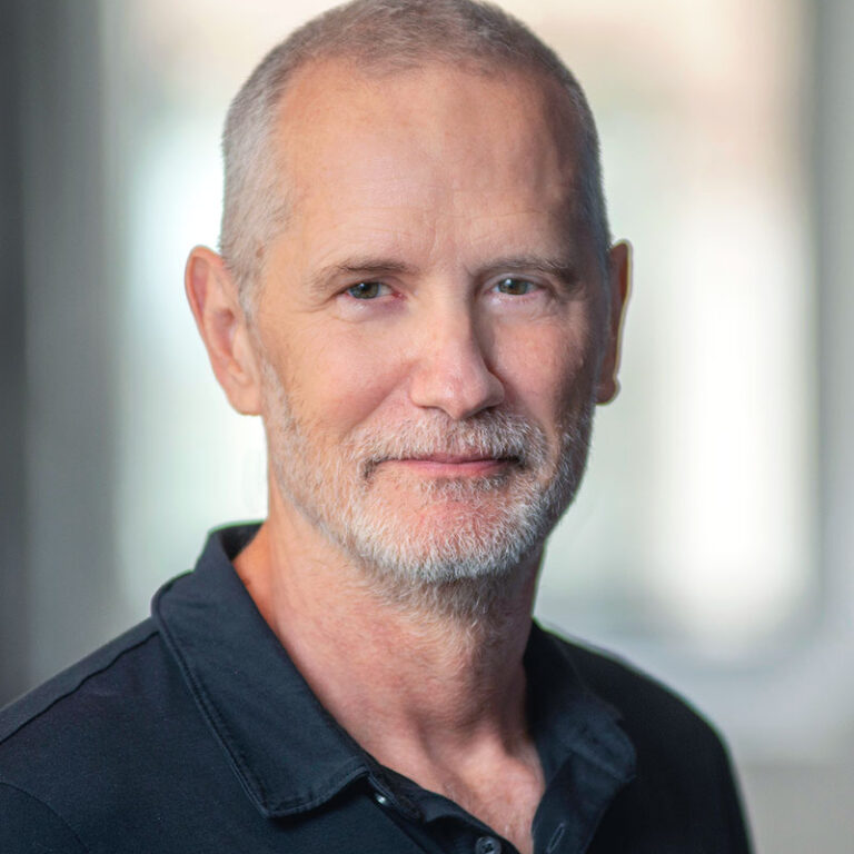 A portrait of a middle-aged man with short hair and a graying beard, wearing a black polo shirt.