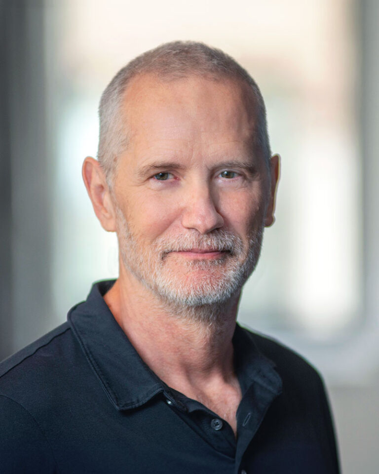 A portrait of a middle-aged man with short hair and a graying beard, wearing a black polo shirt.