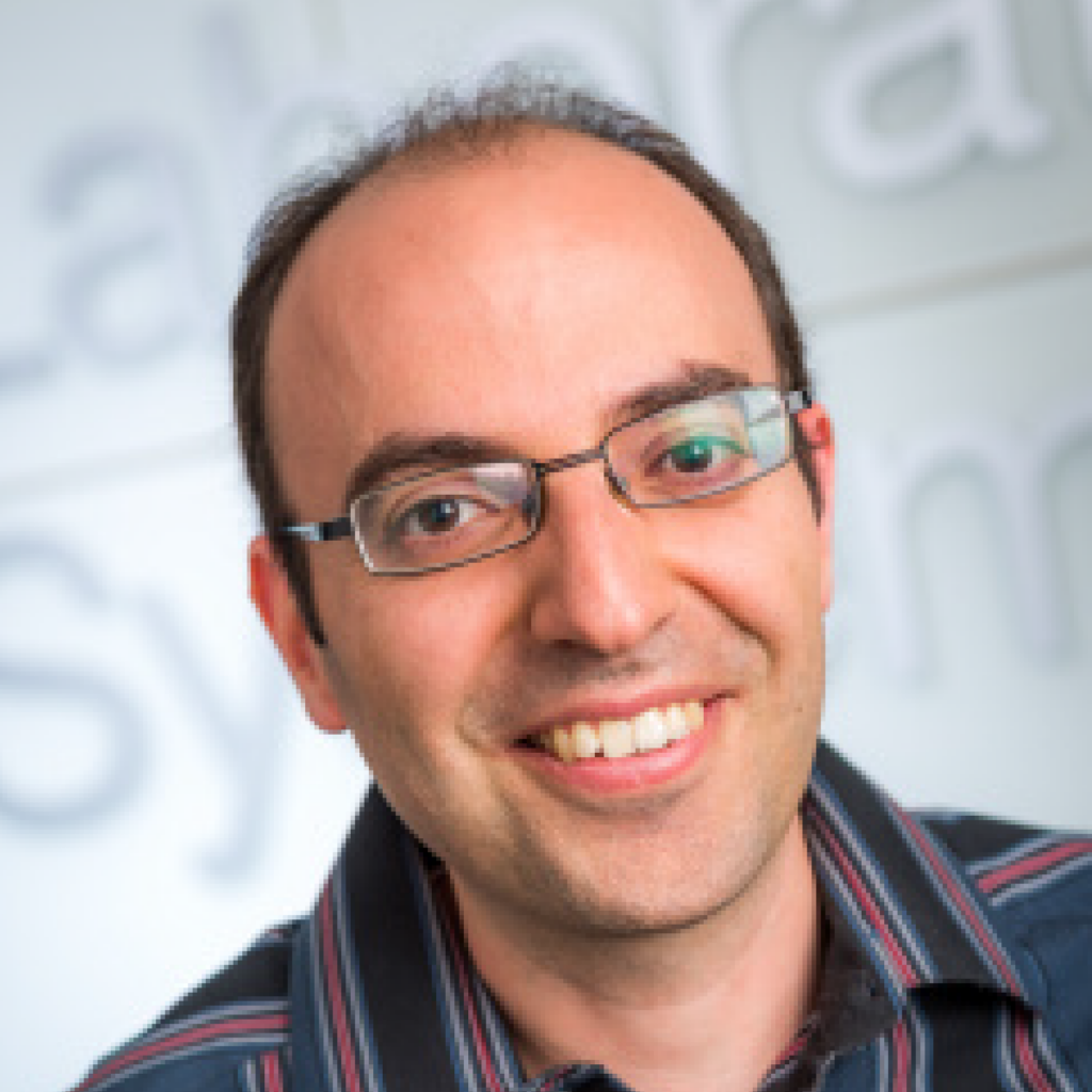 A person with glasses smiles at the camera, wearing a striped shirt. The background is out of focus with some large, indistinct text visible.