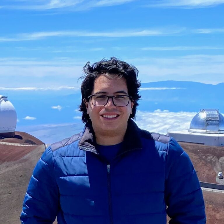 A person wearing glasses and a blue jacket stands outdoors on a mountain with observatory domes and a blue sky in the background.