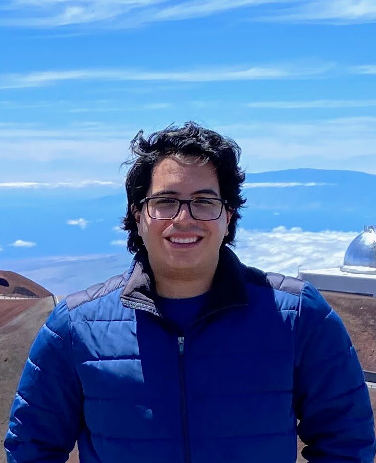 A person wearing glasses and a blue jacket stands outdoors on a mountain with observatory domes and a blue sky in the background.