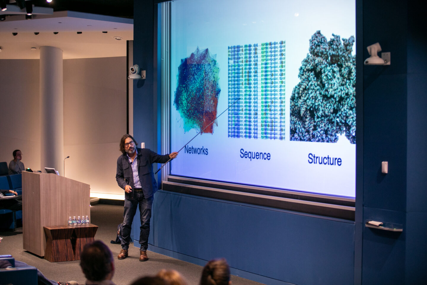 A man presents in a lecture hall, pointing to a projected slide with images labeled Networks, Sequence, and Structure. Audience members are seated and listening.