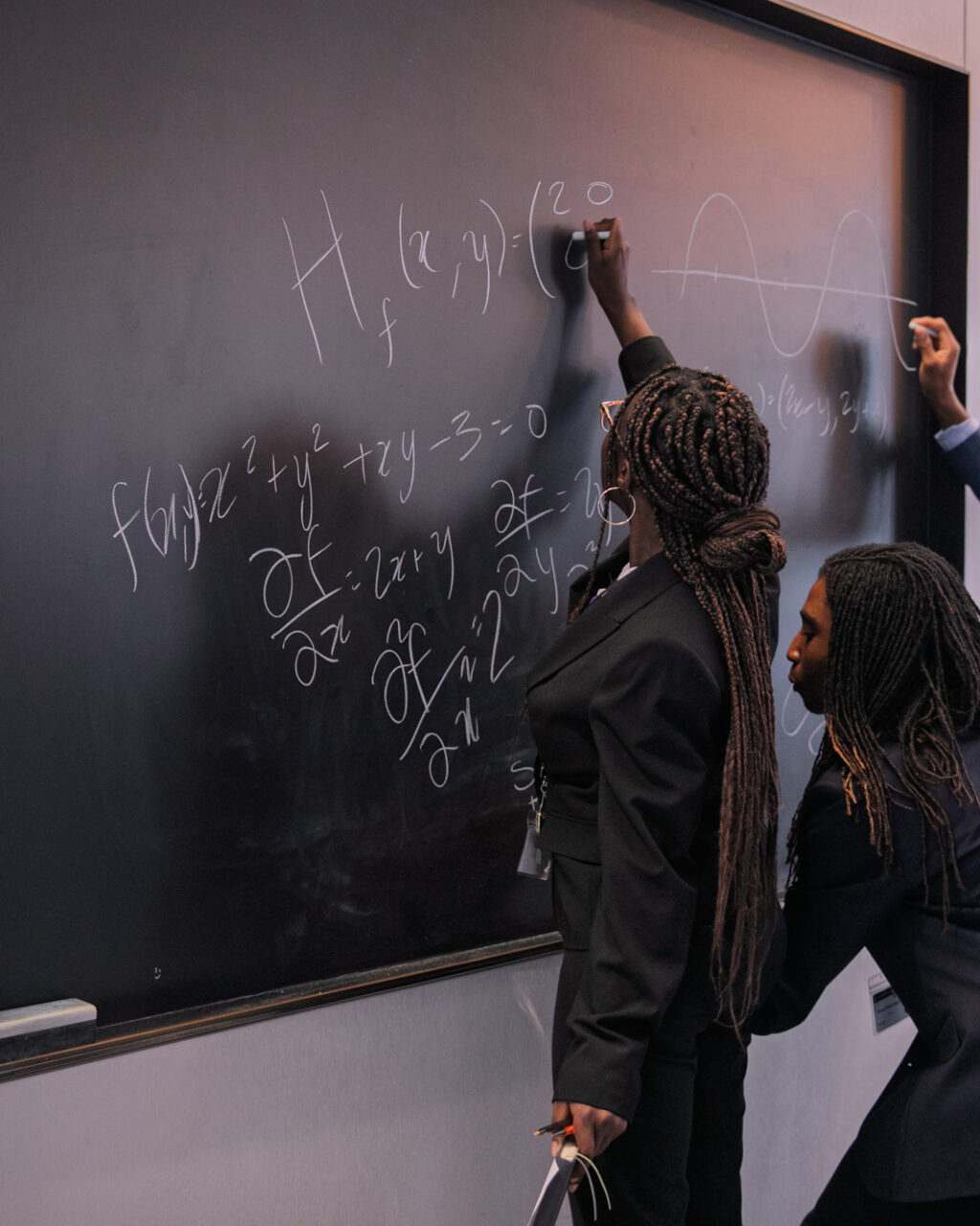 Three people in suits write equations and a wave diagram on a classroom chalkboard. One stands on the left, one crouches to write, and another draws on the right side of the board.