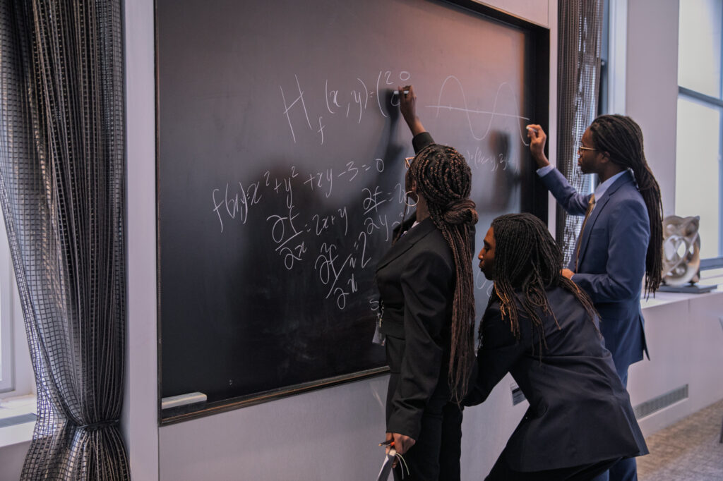 Three people in suits write equations and a wave diagram on a classroom chalkboard. One stands on the left, one crouches to write, and another draws on the right side of the board.
