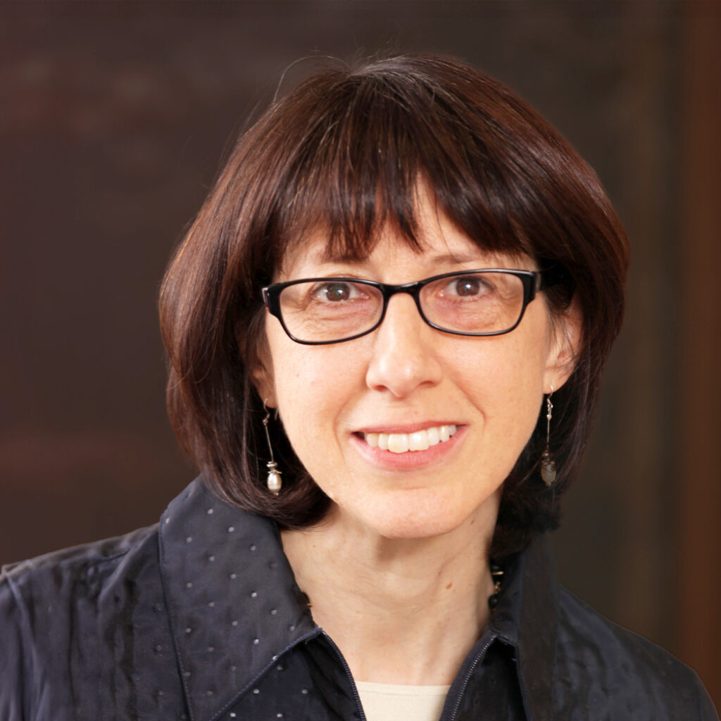 A woman with straight brown hair and glasses smiles at the camera, wearing a dark textured jacket, pearl drop earrings, and a beige top, with a dark blurred background.