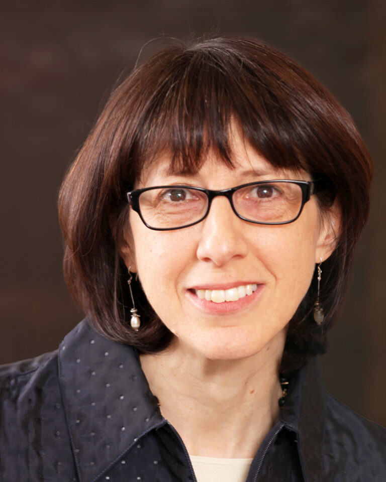 A woman with straight brown hair and glasses smiles at the camera, wearing a dark textured jacket, pearl drop earrings, and a beige top, with a dark blurred background.
