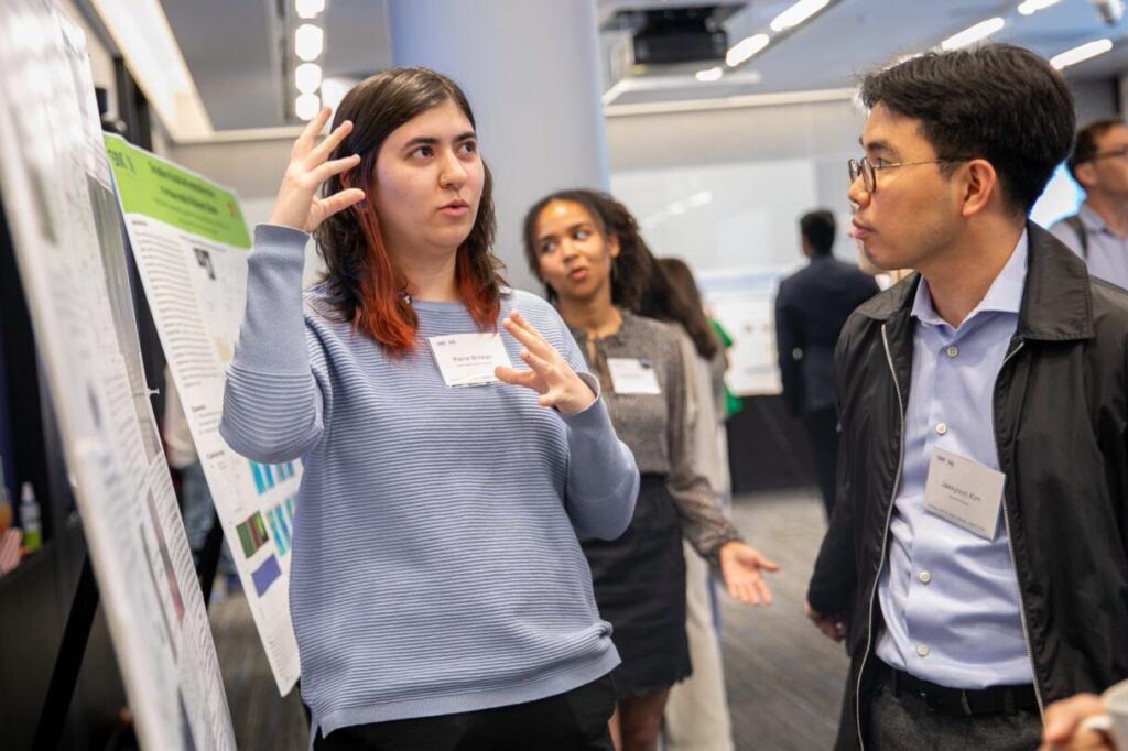 A person gestures while presenting research on a poster to a group at a conference. Attendees listen, and several charts and papers are visible in the background.