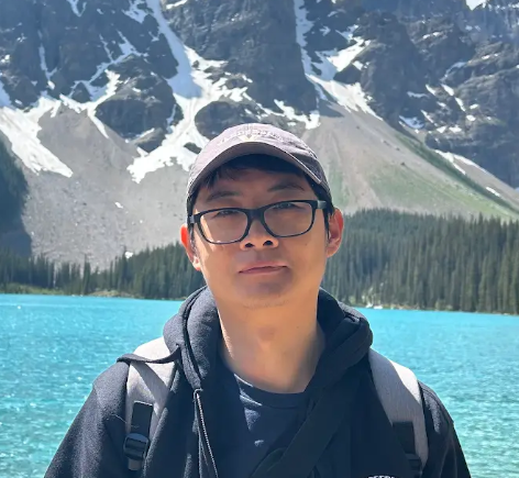 A person wearing glasses, a black cap, and a black hoodie stands by a bright blue lake with pine trees and snow-capped mountains in the background.