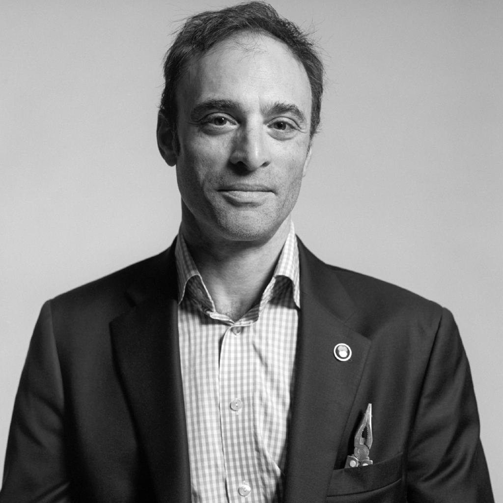 A man in a checked shirt and dark blazer faces the camera, wearing a pin on his jacket and a pen in his pocket, against a plain background.