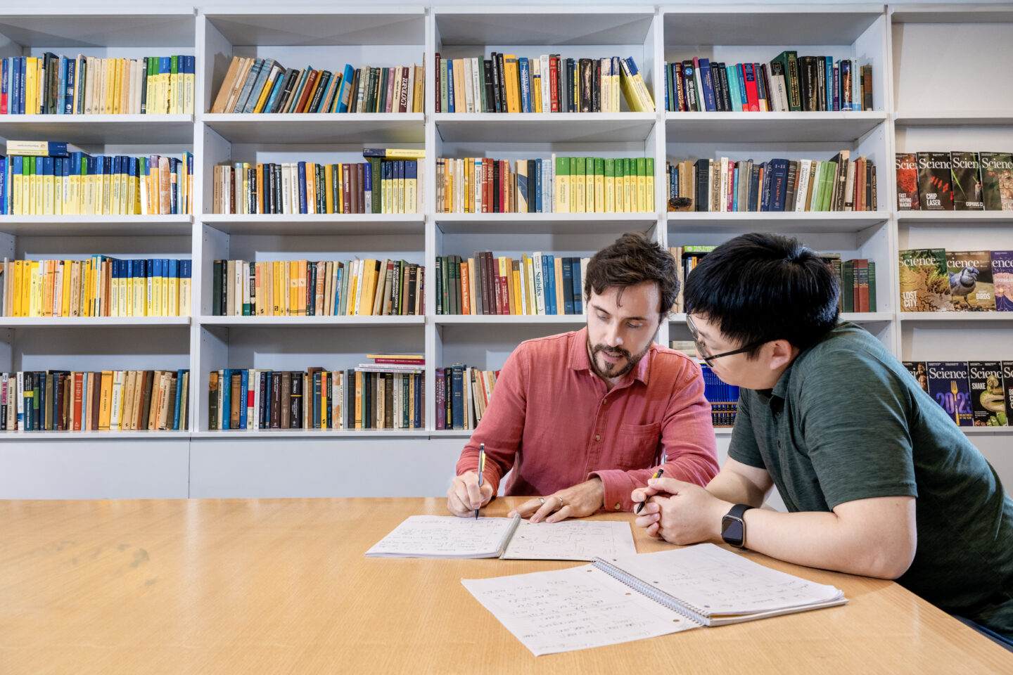 Two people sit at a table in a library, writing and discussing notes, with open notebooks and a bookshelf filled with colorful books in the background.