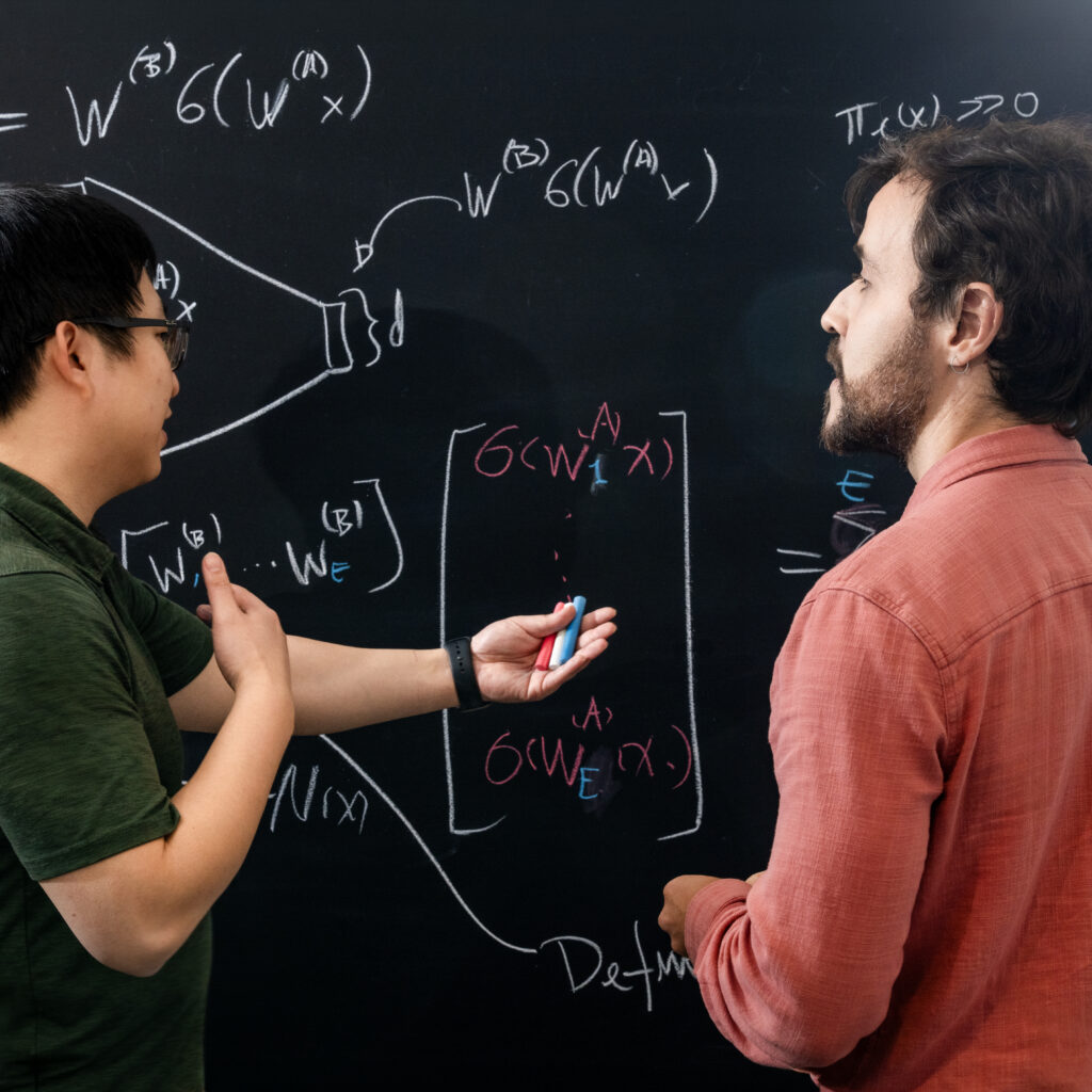 Two people stand in front of a blackboard covered with complex mathematical equations, discussing and pointing at the formulas while holding colored chalk.