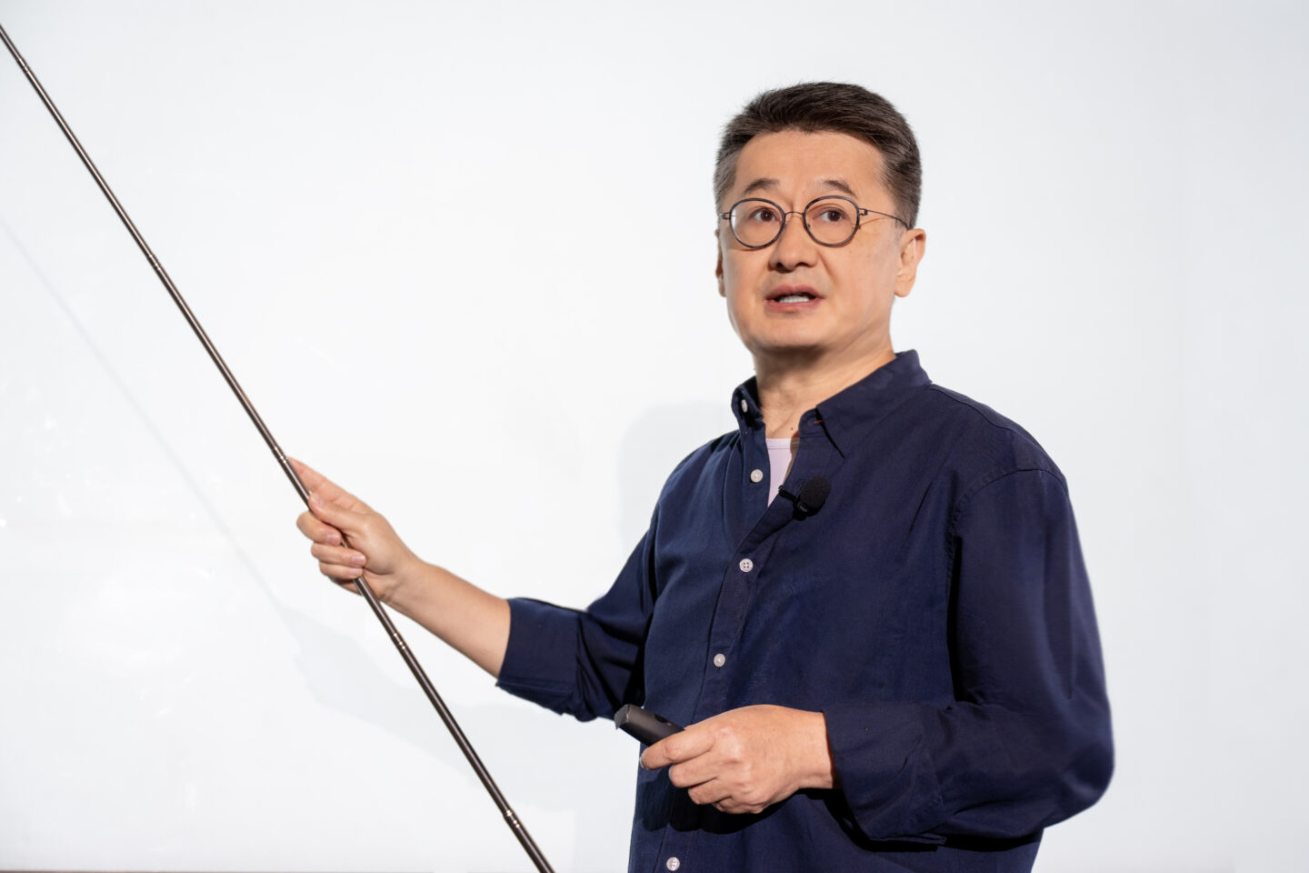 A person wearing glasses and a dark shirt points at a blank whiteboard with a pointer, while holding a marker. They appear to be giving a lecture or presentation.