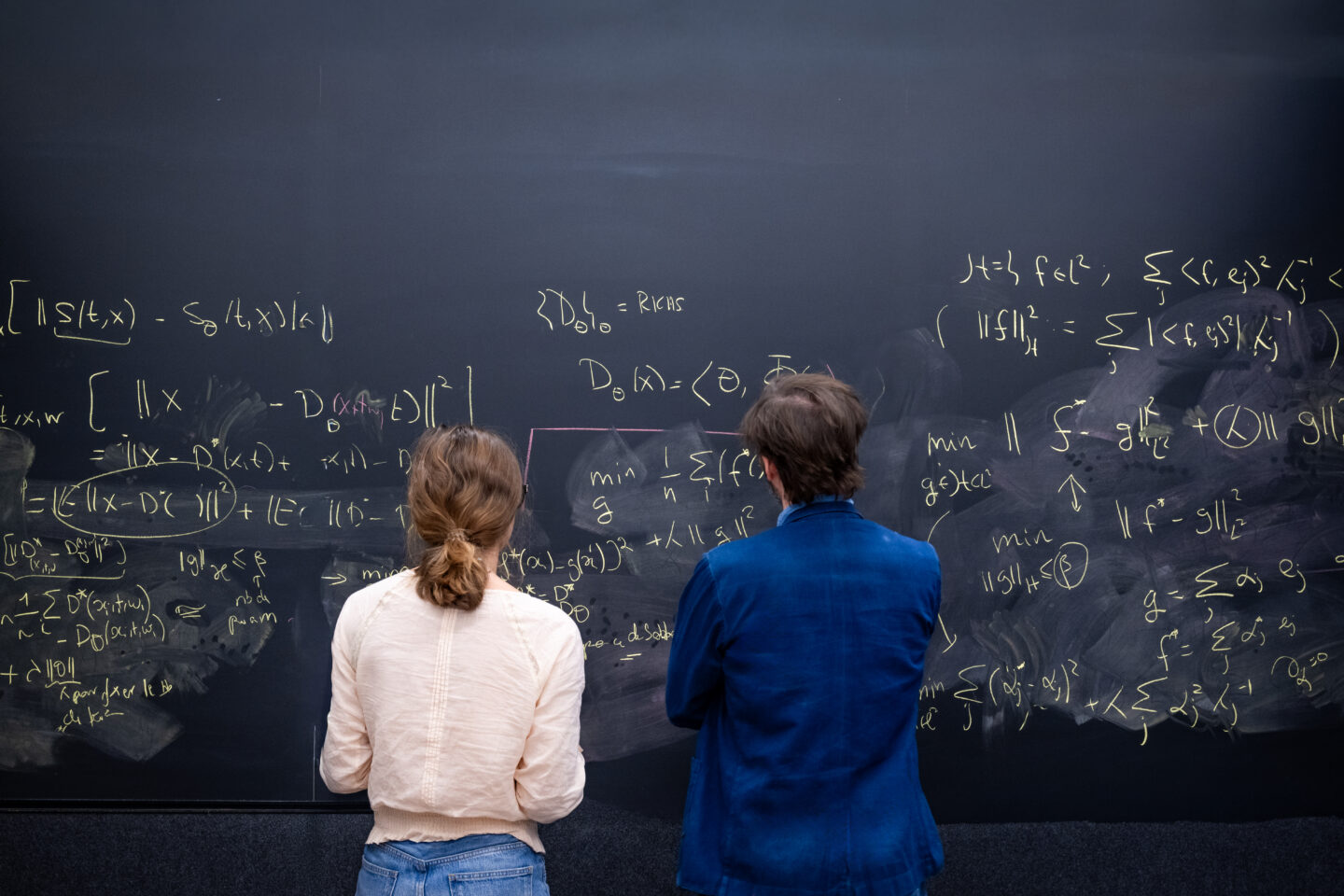 Two people stand in front of a large blackboard filled with mathematical equations written in yellow chalk, appearing to discuss and analyze the work together.