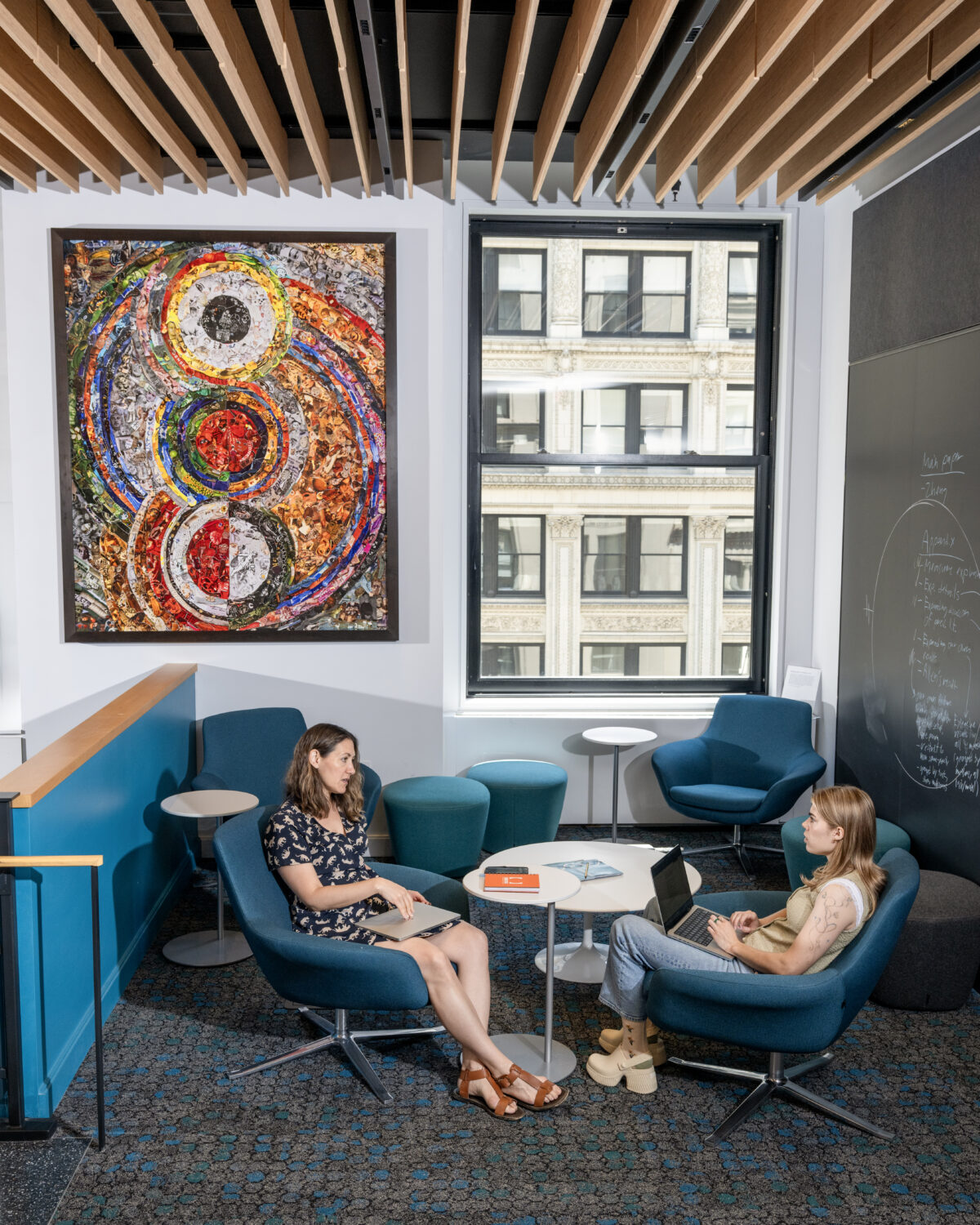 Two women sit in a modern office lounge, talking and working on laptops. A colorful abstract painting hangs on the wall, and large windows reveal a city building outside. A round table is between them.