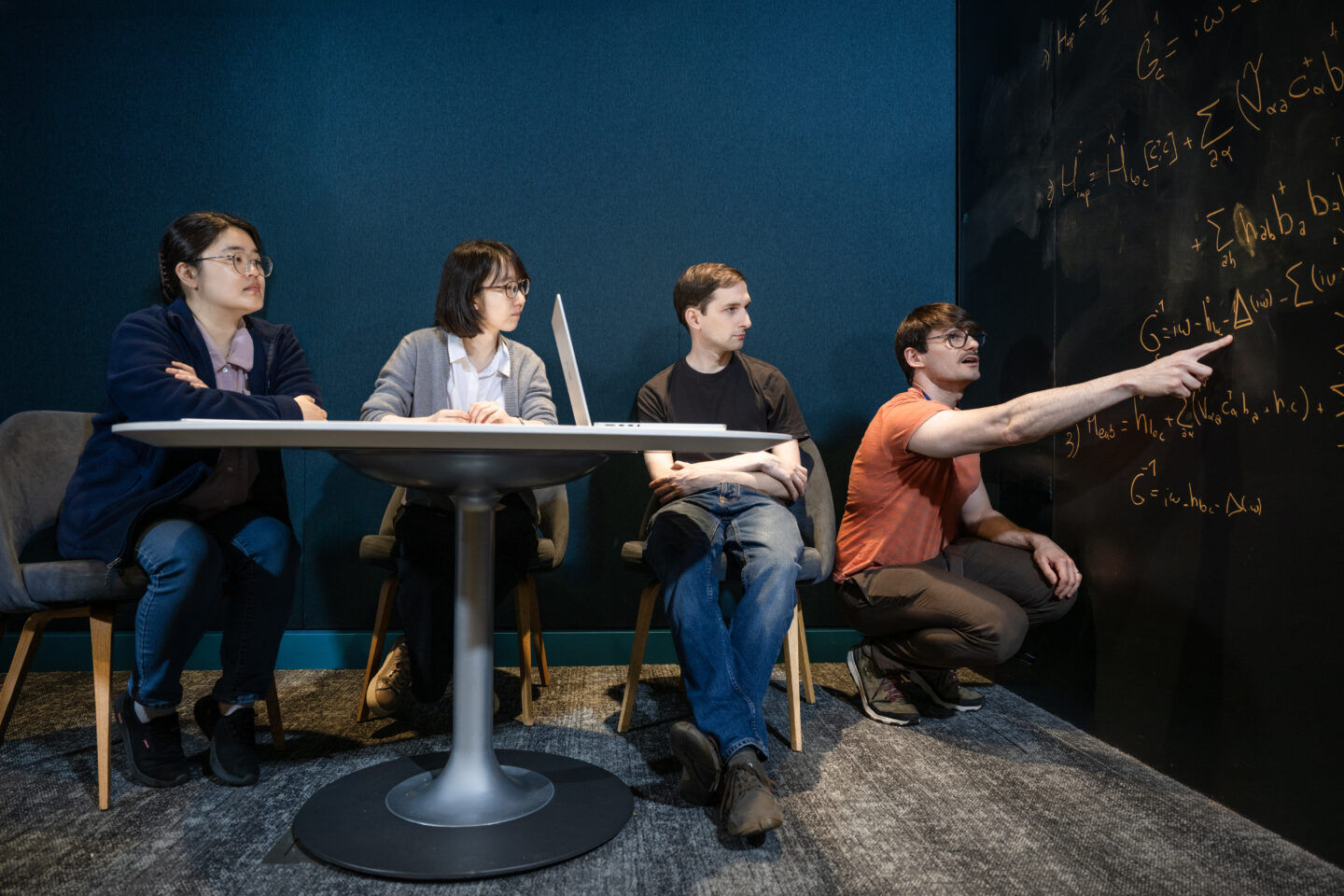 Four people sit around a table, attentively watching a person kneeling and pointing at complex equations written on a large black wall in a modern, well-lit room.